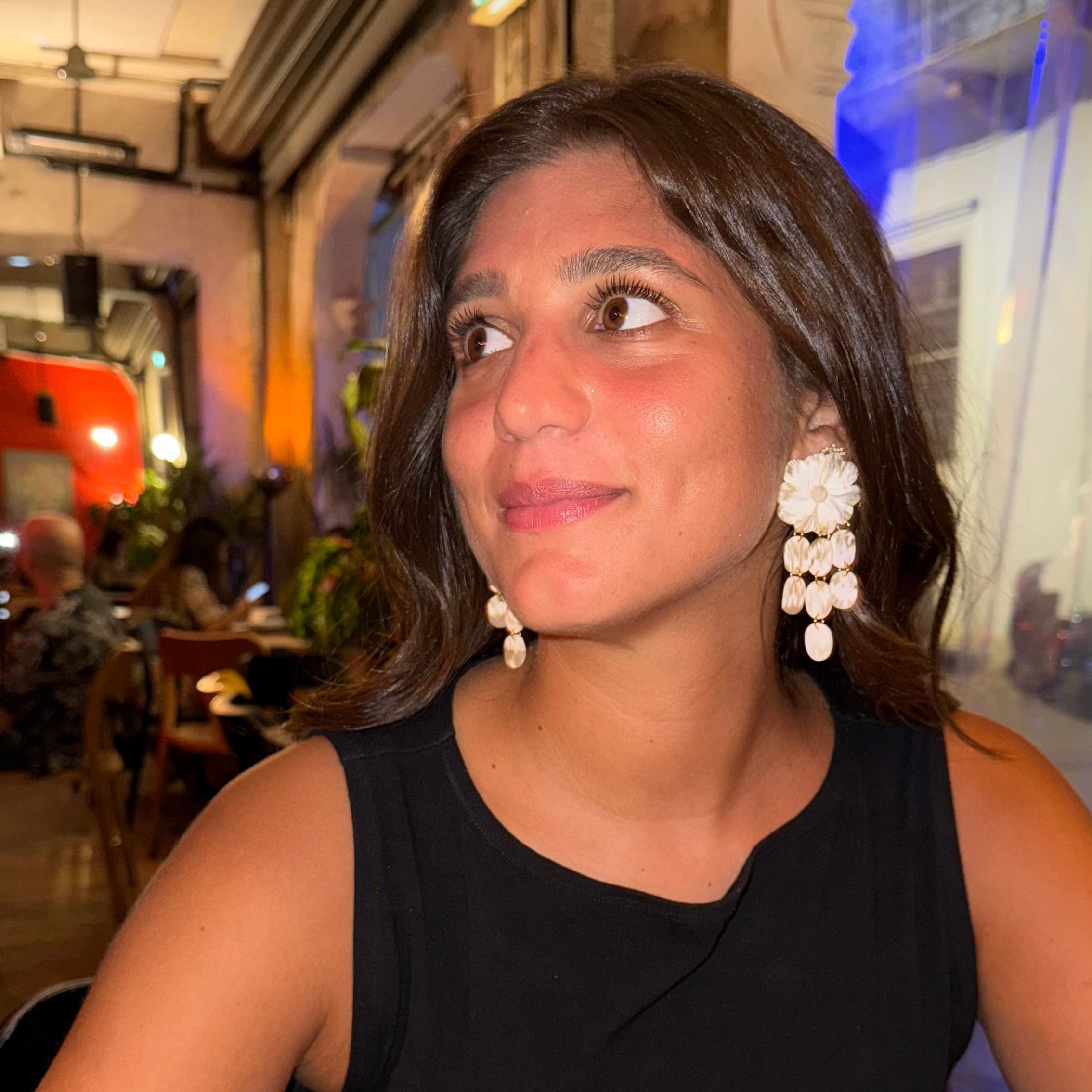Woman sitting at a table in an indoor setting, wearing a black sleeveless top and white earrings.