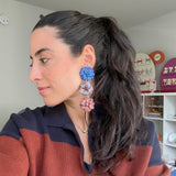 A woman with dark hair in a ponytail is shown in profile wearing a long, dangling floral earring with blue, white, and pink flower charms. She is wearing a rust and navy striped knit top, and the background shows a home interior with shelves.