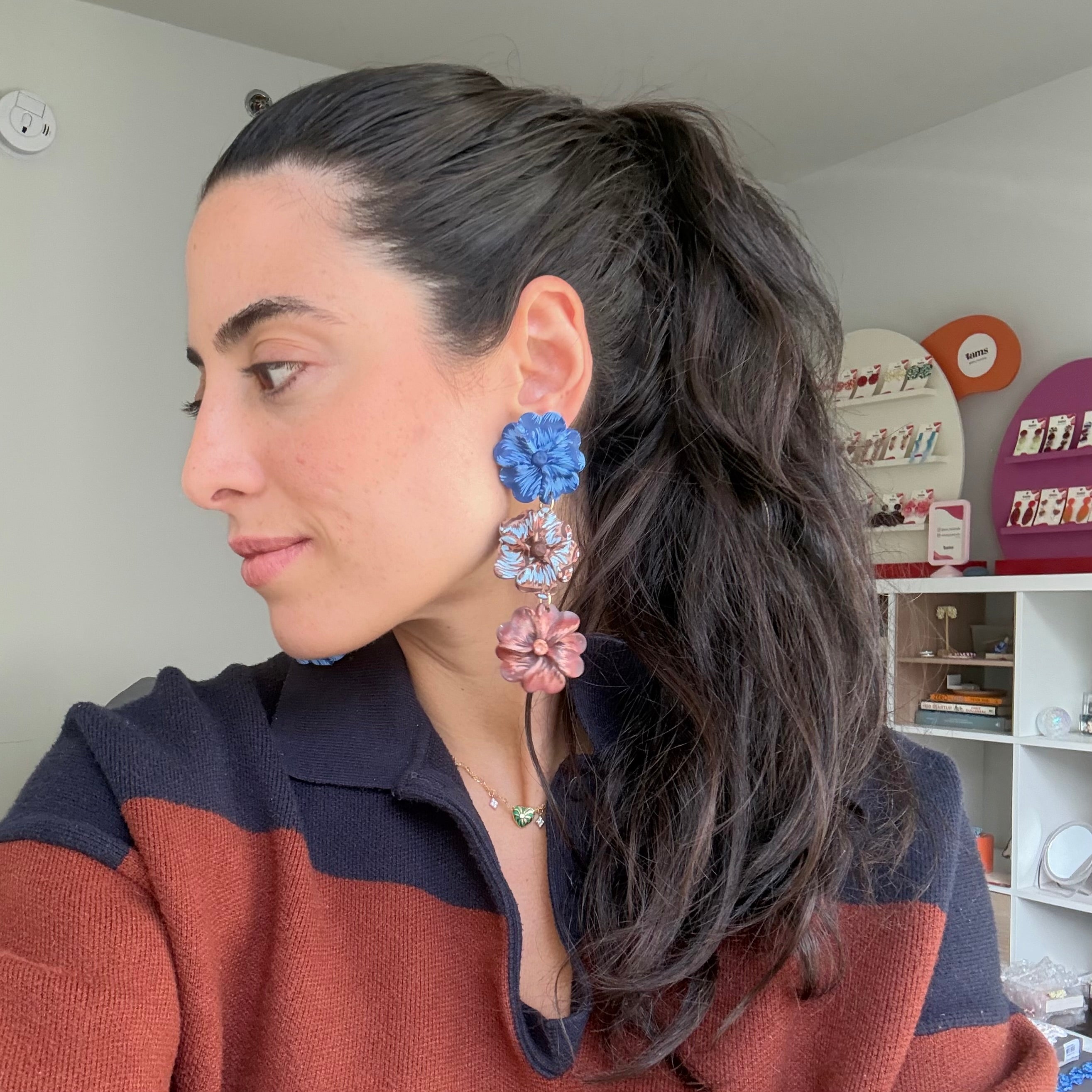 A woman with dark hair in a ponytail is shown in profile wearing a long, dangling floral earring with blue, white, and pink flower charms. She is wearing a rust and navy striped knit top, and the background shows a home interior with shelves.