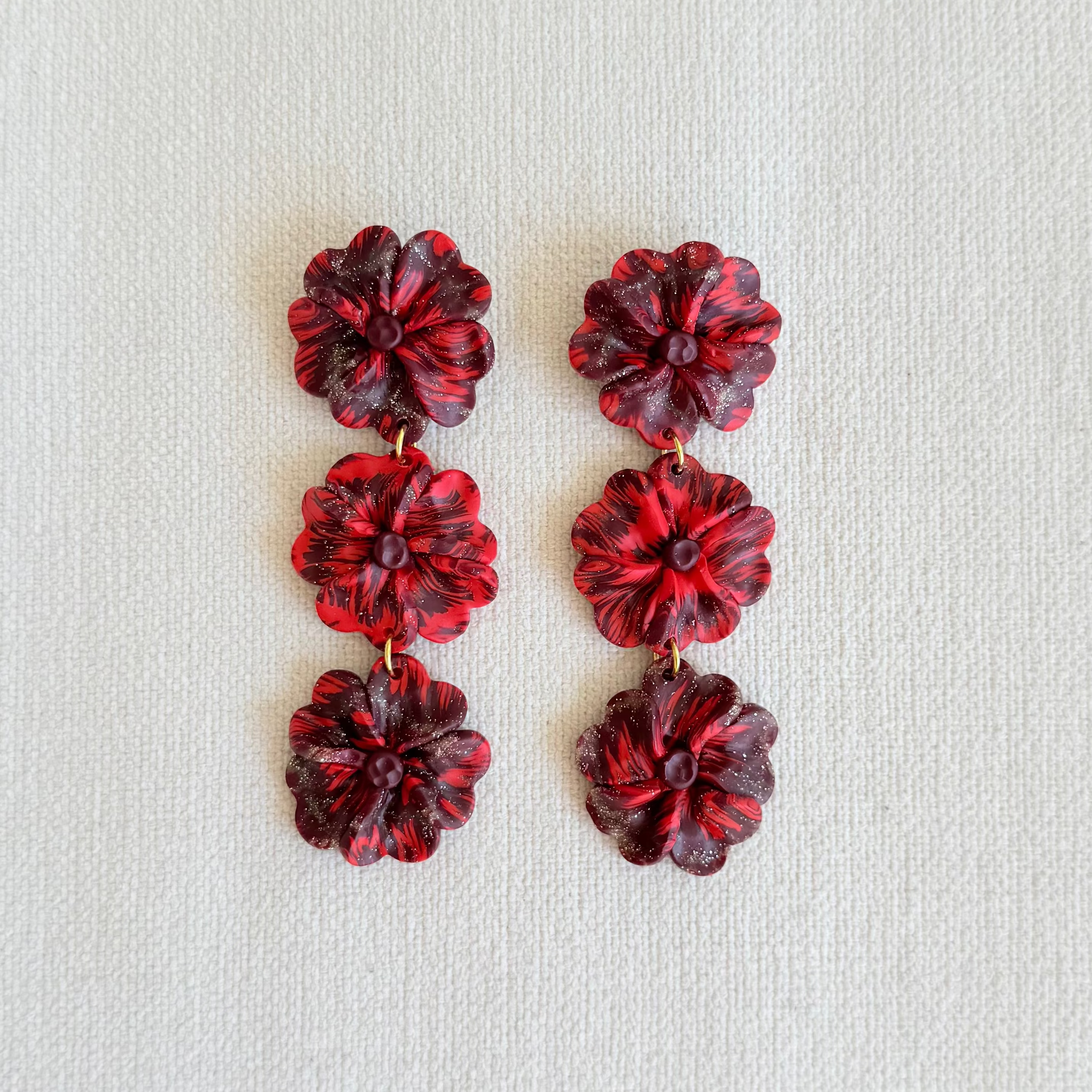 A pair of red, burgundy and glittery marbled flower-shaped drop earrings arranged vertically on a textured off-white fabric background.