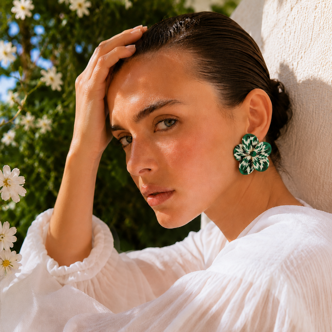 A woman in a white blouse wearing a large green and white floral statement earring, posing outdoors against a textured wall with white flowers and green foliage in the background.